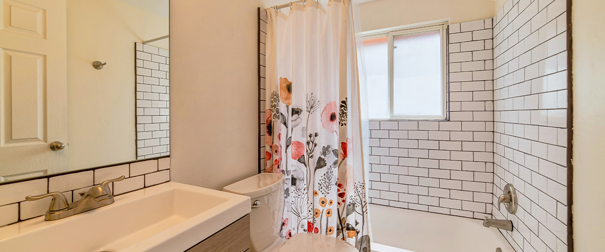 A cozy bathroom featuring a floral shower curtain, white subway tiles, a vanity with a large mirror, and modern fixtures.